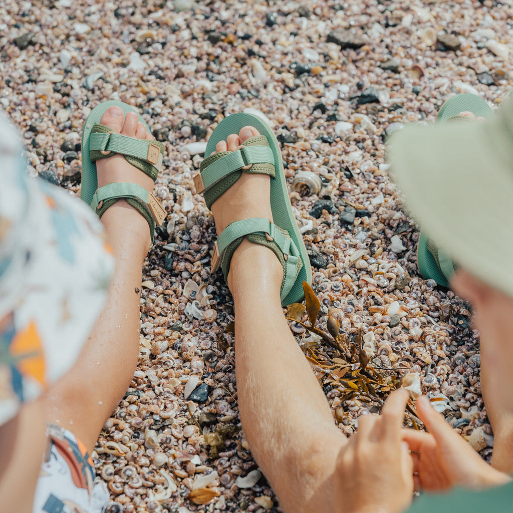 Two children sitting on a pebble beach wearing CRYWOLF beach sandals. 